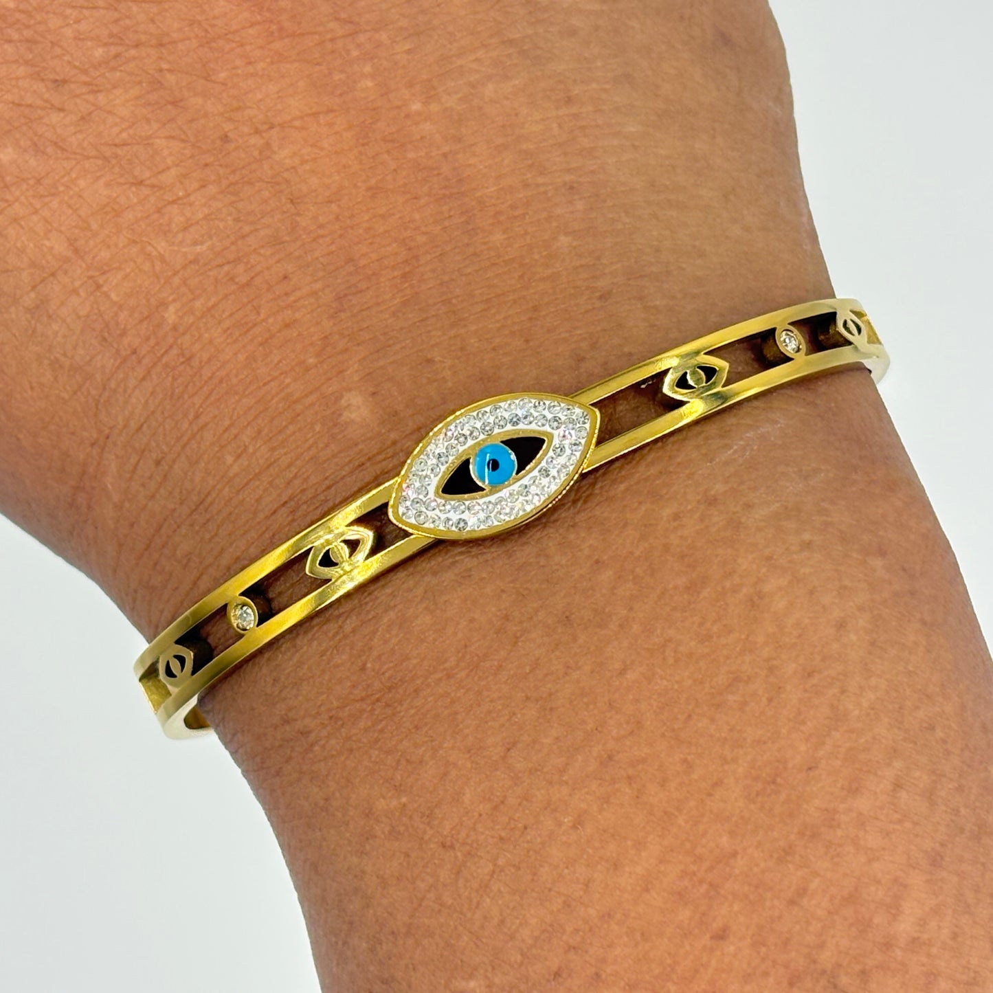 A gold-toned metal cuff bracelet with an evil eye charm in the center, worn on a person's wrist. The evil eye charm features a blue center and is surrounded by a halo of small clear stones.