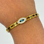 A gold-toned metal cuff bracelet with an evil eye charm in the center, worn on a person's wrist. The evil eye charm features a blue center and is surrounded by a halo of small clear stones.